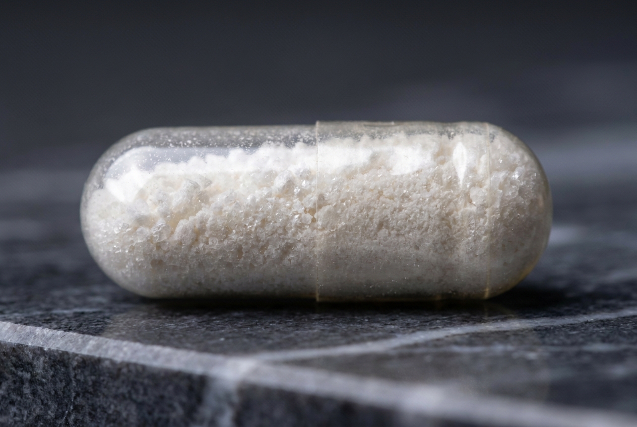 Close-up of a clear supplement capsule filled with white powder on a dark marble surface.